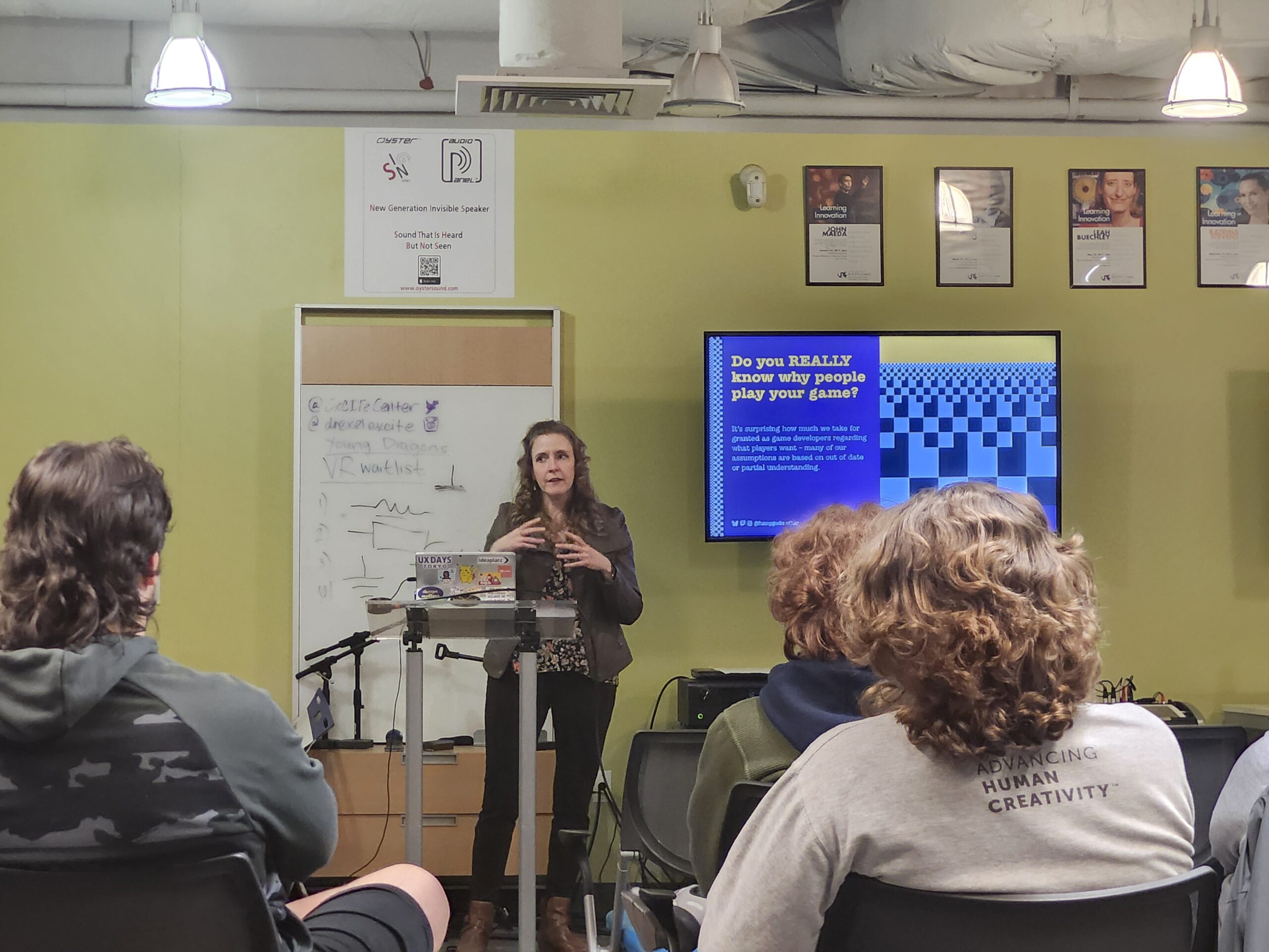 Author Cheryl Platz stands in front of a slide depicting the nine Motivators of Play from her book The Game Development Strategy Guide while giving a guest lecture at Drexel University's Entrepreneurial Game Studio on November 5, 2025. Three students are seen in the foreground.