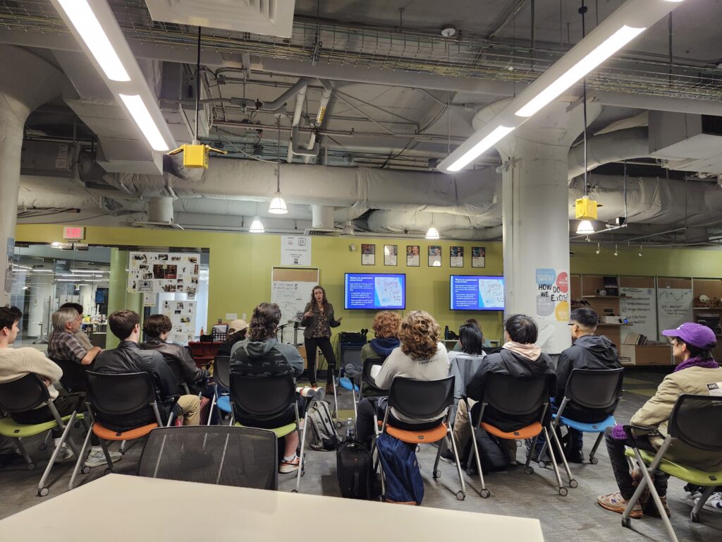 Author Cheryl Platz stands in front of a slide depicting Raph Koster's Theory of Fun while providing background on the Motivators of Play from her book The Game Development Strategy Guide while giving a guest lecture at Drexel University's Entrepreneurial Game Studio on November 5, 2025. A number of students and instructors are seen in the foreground.