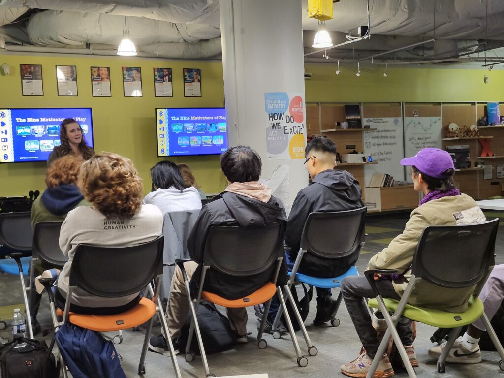 Author Cheryl Platz stands in front of a slide depicting the nine Motivators of Play from her book The Game Development Strategy Guide while giving a guest lecture at Drexel University's Entrepreneurial Game Studio on November 5, 2025. A number of students and instructors are seen in the foreground.