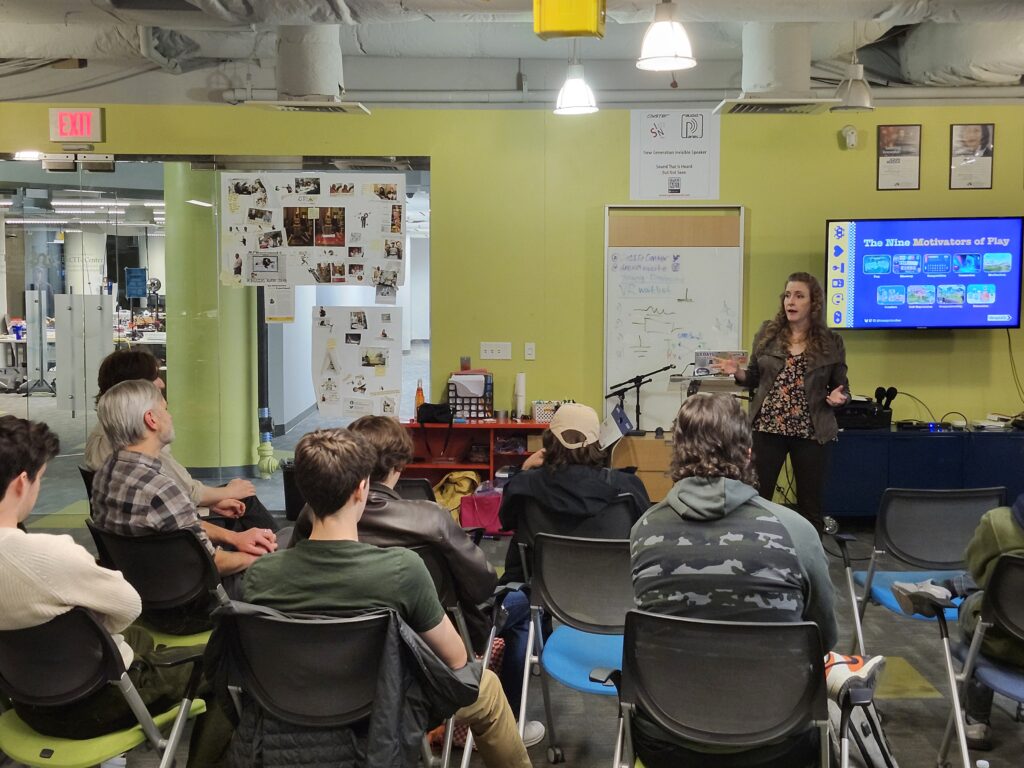 Author Cheryl Platz stands in front of a slide depicting the nine Motivators of Play from her book The Game Development Strategy Guide while giving a guest lecture at Drexel University's Entrepreneurial Game Studio on November 5, 2025. A number of students and instructors are seen in the foreground.